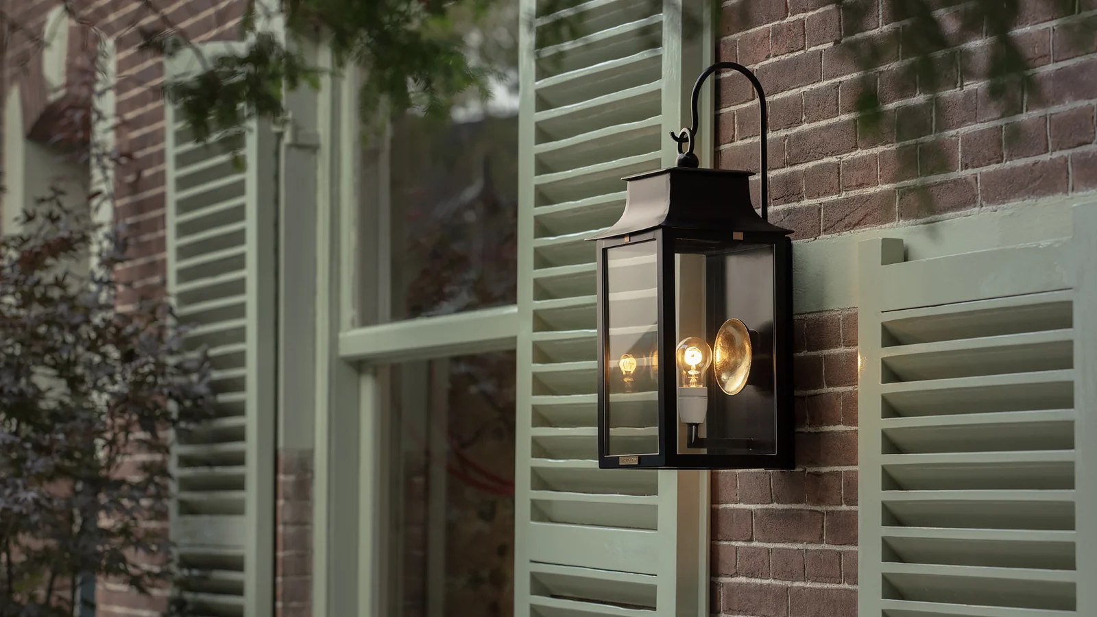 Een Artisans-buitenlamp met warm licht, aan een gevel met groene luiken bij Villa Arte.