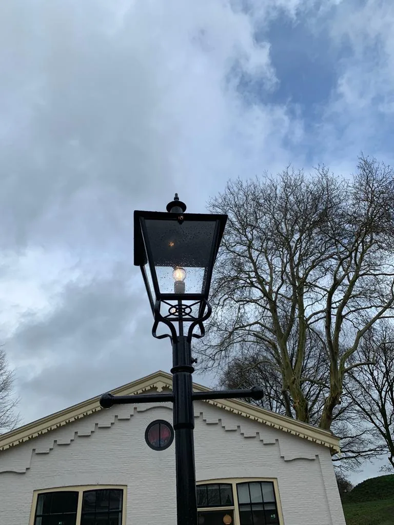 Zwarte lantaarnpaal voor een witte gevel met bomen op de achtergrond.